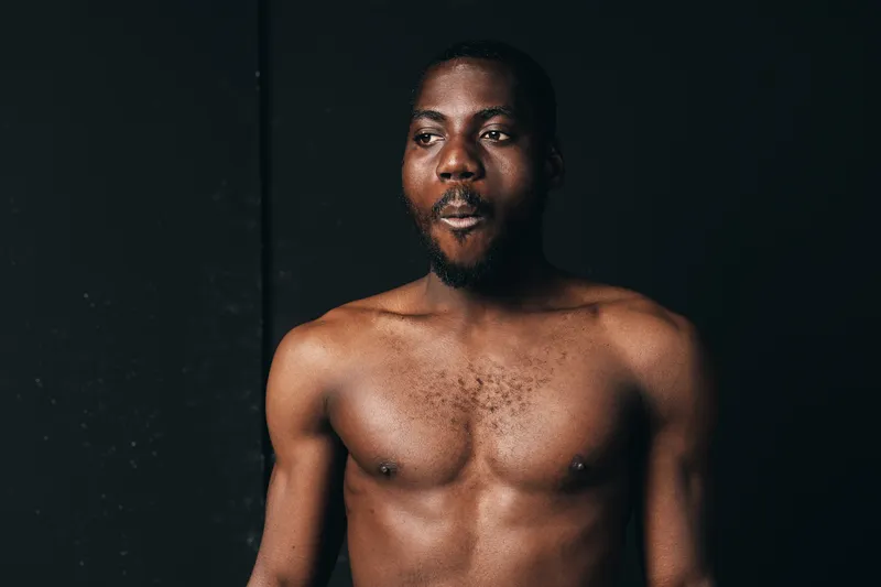 African man shirtless with muscular body and beard in creative conceptual visual style, dark background, portrait with serious expression and natural lighting.