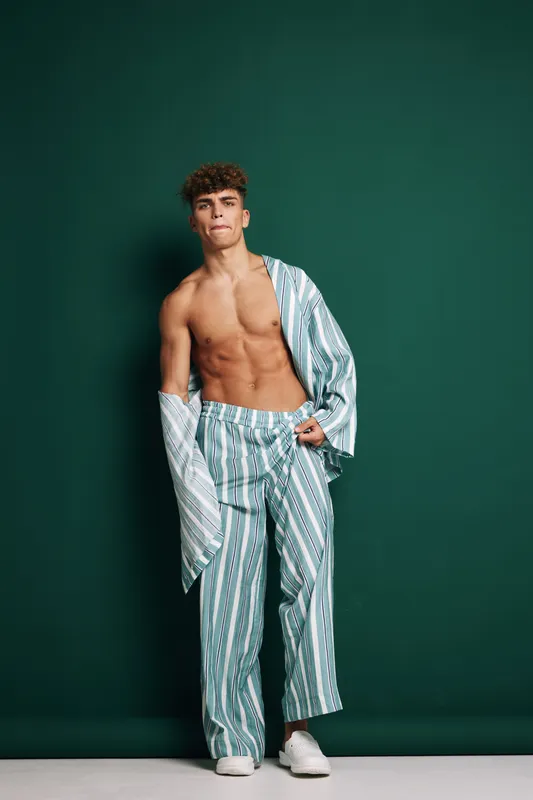 Young man with curly hair posing shirtless wearing striped pajama pants and open pajama shirt against green background in studio, casual and relaxed fashion style portrait.