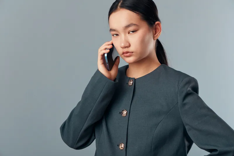 Young Asian woman with serious expression talking on phone, wearing dark jacket with gold buttons, studio portrait on gray background with focused and professional mood.