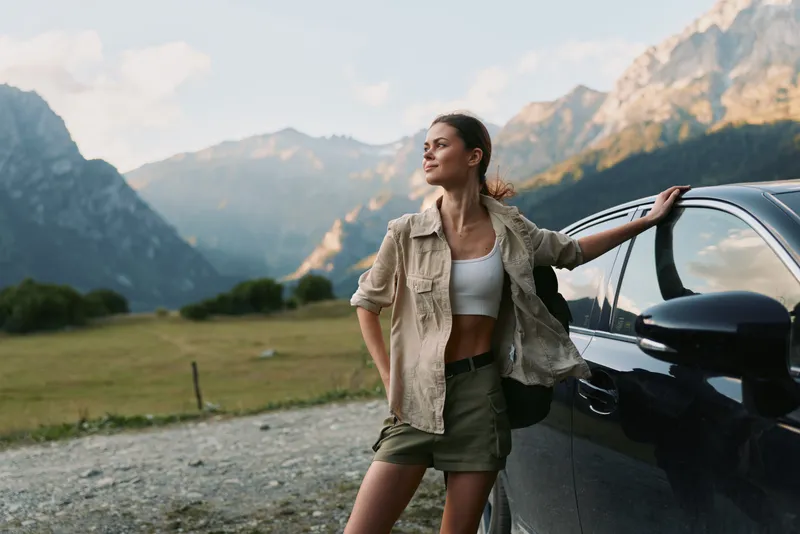 A solo traveler, a young woman, leans on a dark car in a mountain landscape, wearing a white crop top, khaki shorts and a beige jacket, exuding casual adventure.