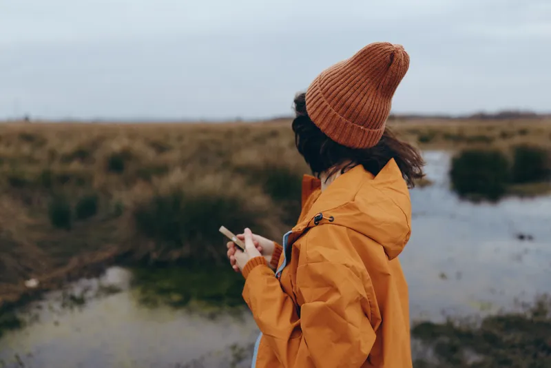 Woman lifestyle outdoors in winter countryside, standing by river, wearing orange coat and beanie, holding smartphone and taking photo, calm portrait with soft cloudy sky and fields