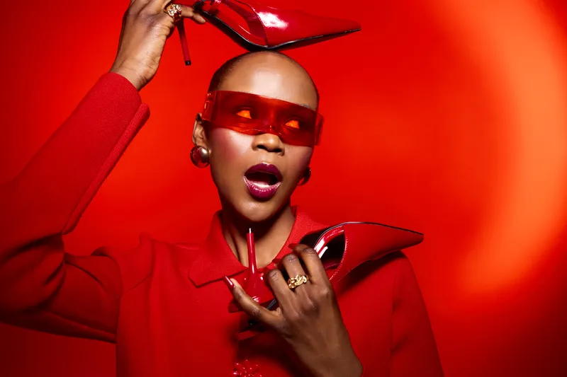 Stylish dark skinned African woman in red coat and sunglasses holds red high heels near face on red background. Concept of fashion, glamour, bold style, and confident beauty portrait.