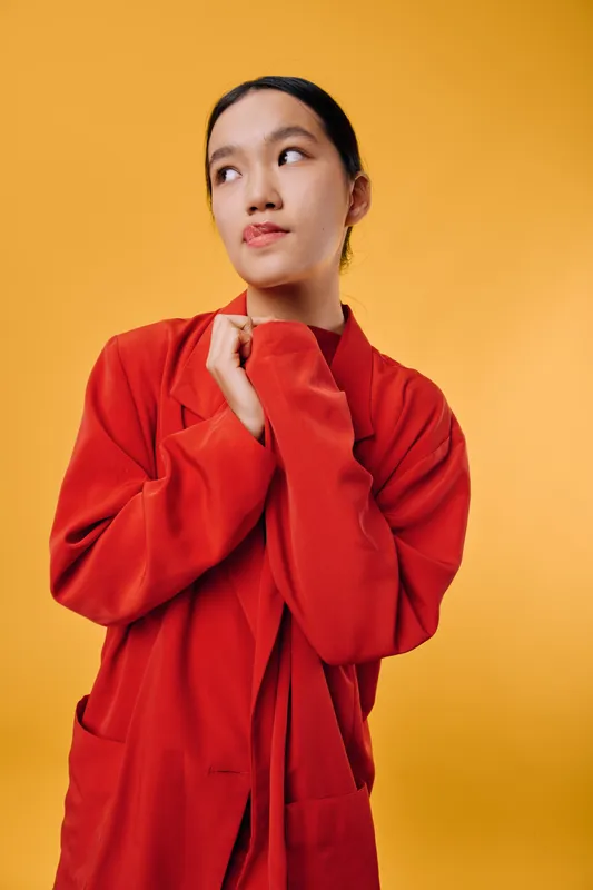 Young woman in red coat looking sideways with thoughtful expression against plain yellow background. Portrait of confident female with black hair wearing casual outerwear indoors.