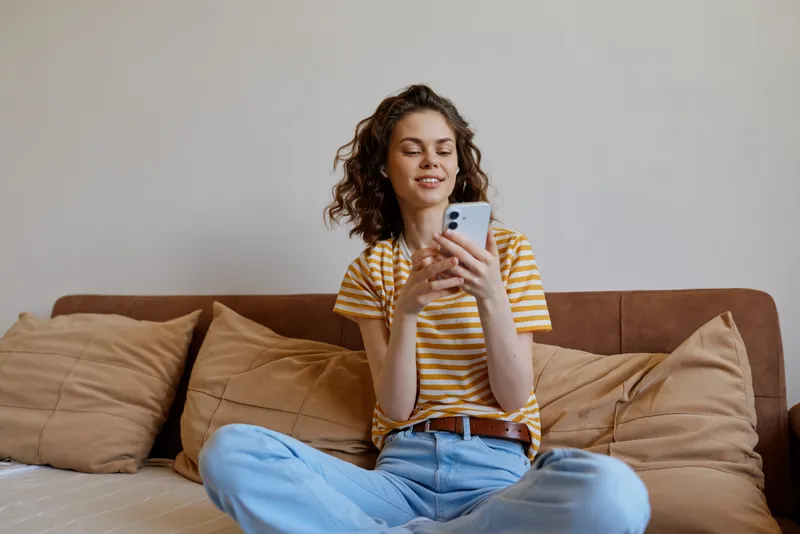Cheerful woman indoors, casual lifestyle moment, striped shirt, sitting on couch, using smartphone, warm natural light, cozy home setting, relaxed vibe