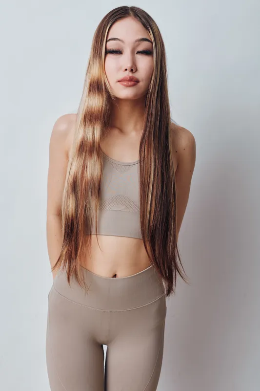 Young woman with long straight hair and closed eyes, wearing beige sport top and leggings standing against a plain background in a relaxed posture for fashion or wellness.