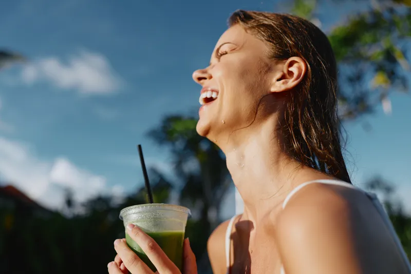 Cheerful woman outdoors, sipping a green smoothie, sunlit moment, carefree smile, breezy casual vibe, vibrant colors, lush trees in the background