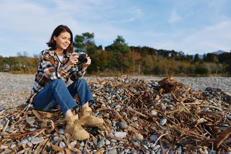 Woman photographer sitting on a rocky shore in autumn scenery, capturing moments with a handheld camera near a calm lake, colorful trees and open sky.