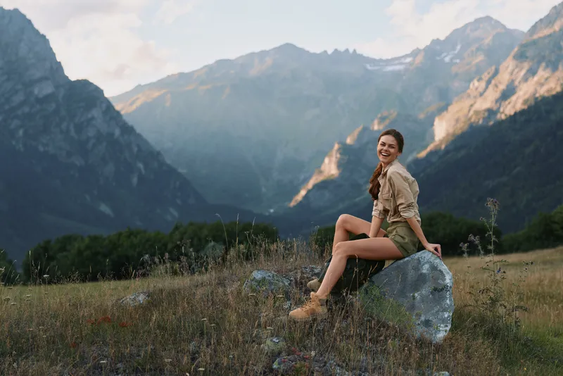 A woman sits on a rock in a mountain meadow, enjoying the scenery. Bright natural light highlights her relaxed pose and outdoor lifestyle.