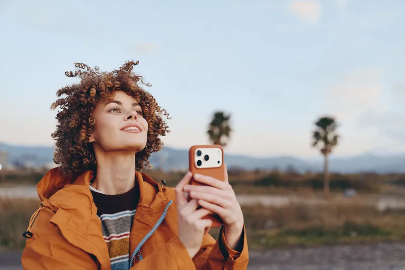 Curly hair woman in orange jacket takes a selfie outdoors, smiling while holding a smartphone, lifestyle portrait in golden hour, blurred field landscape, communication and connection
