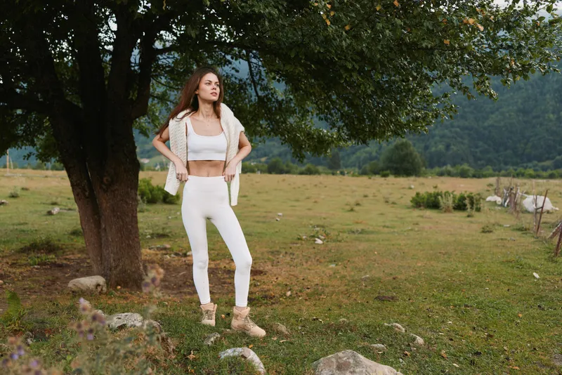 Young woman in a white crop top and pants stands beneath a leafy tree in a sunlit meadow, hands on hips, exuding relaxed confidence and casual countryside style.