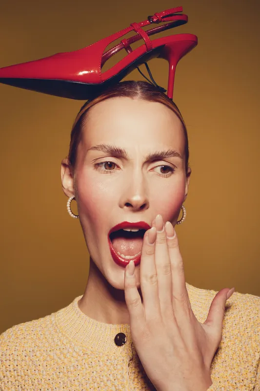 Surprised woman with red lipstick and hoop earrings holds hand near mouth. A red high heel shoe is balanced on her head against a warm brown background. Concept of fashion and expression.