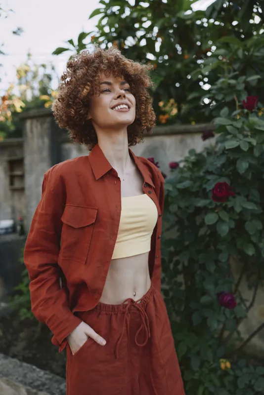 Joyful woman outdoors, wearing red jacket and pants with yellow crop top, casual lifestyle portrait in a sunlit garden, natural warm smile