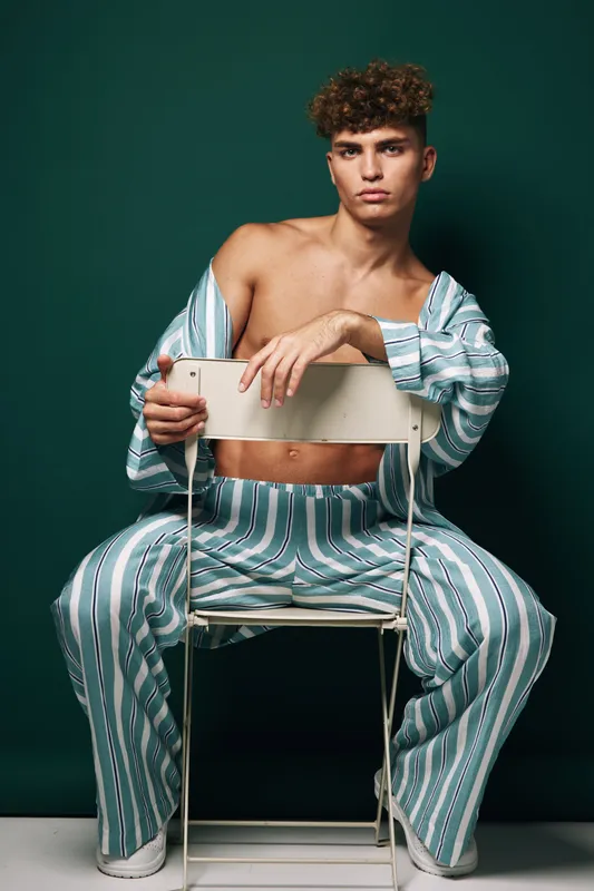 Young man with curly hair sitting on chair backwards wearing striped pants and open shirt against green background in studio portrait.