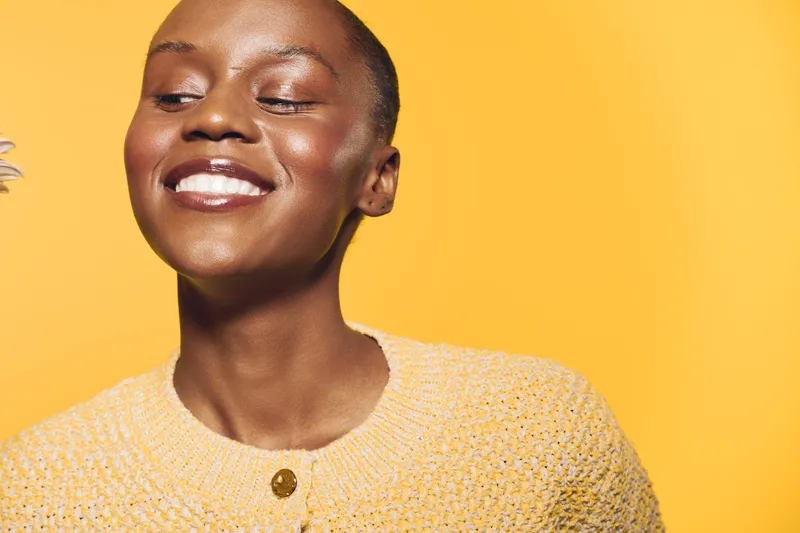Happy African woman with dark skin smiles with closed eyes wearing yellow knitted sweater. Bright yellow background creates warm and joyful mood. Concept of beauty, happiness, and wellness.