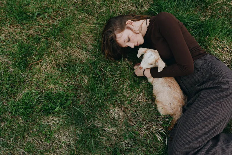 A woman lies on grass, cuddling a small goat kid in a peaceful outdoor moment. Warm natural light highlights tenderness, nature, lifestyle, and cozy affection.