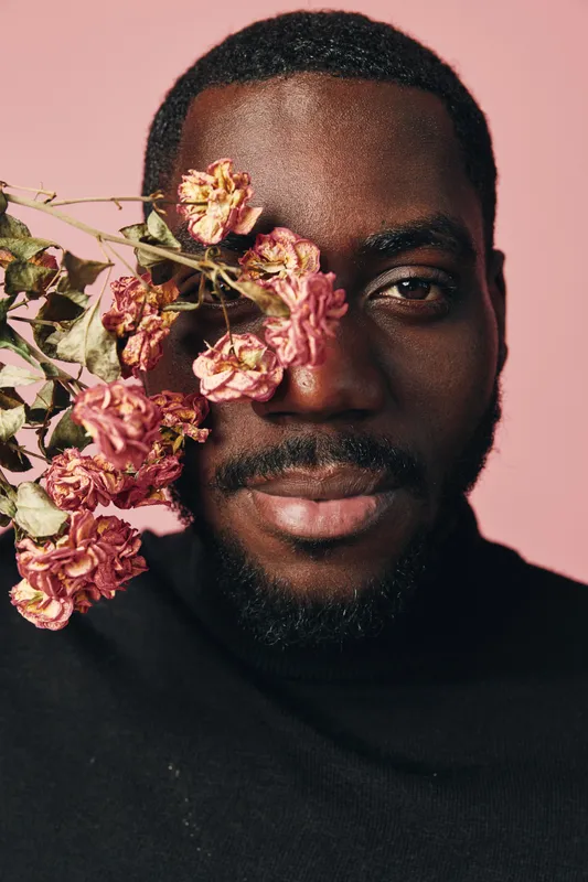 Creative portrait of African man with dried pink flowers covering one eye, wearing black turtleneck against pastel pink background in conceptual visual style.