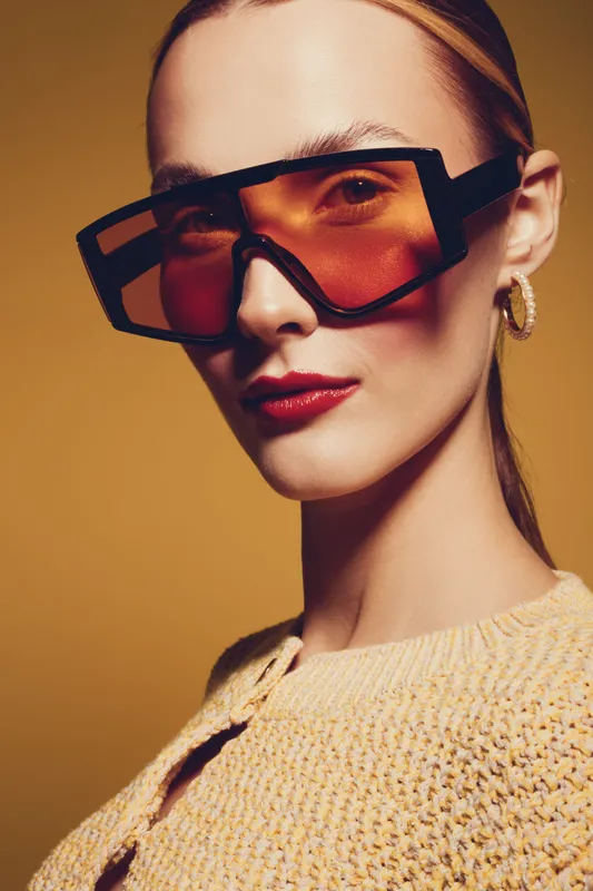 Stylish woman with oversized orange sunglasses and hoop earrings poses confidently against a warm brown background. Fashion portrait with modern accessories and makeup concept.