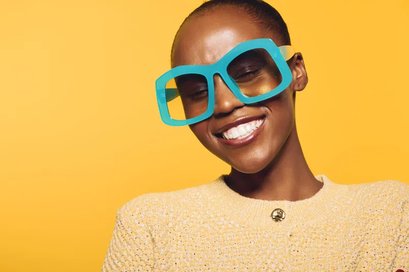 Smiling dark skinned African woman wears oversized blue glasses and yellow sweater, posing against yellow background. Concept of fashion, joy, and confident black female model portrait.