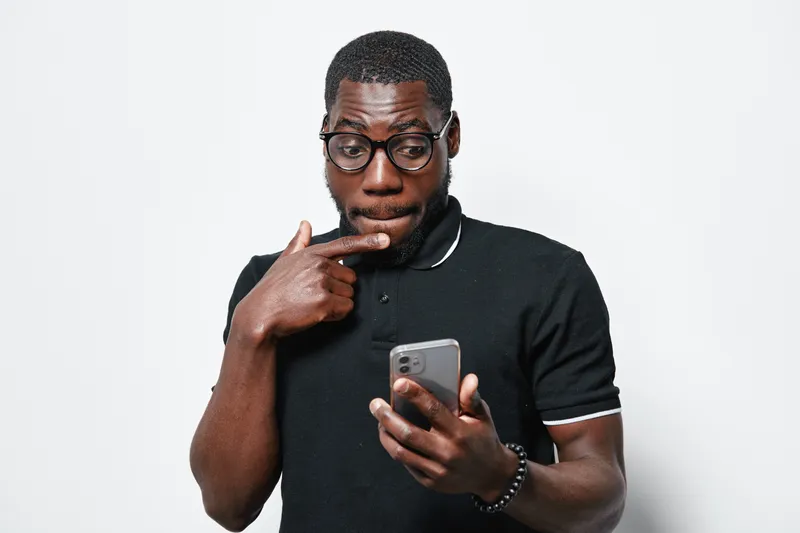 African American man with dark skin and glasses looking surprised at smartphone screen wearing black polo shirt on white background. Casual technology use and facial expression.