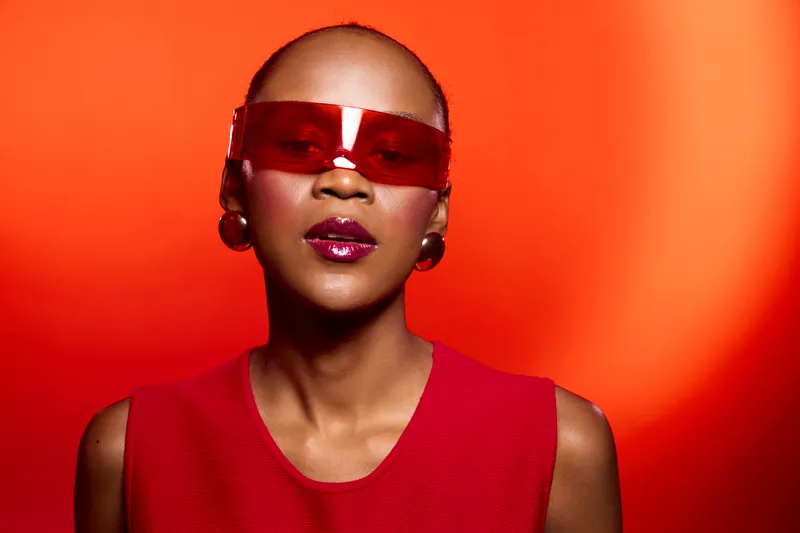 Stylish dark skinned African American woman wearing large red sunglasses and red sleeveless top poses confidently against vibrant red background. Concept of fashion, bold accessories, and beauty.