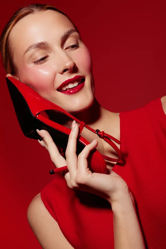 Young woman in red dress holds red high heel shoe near face with closed eyes and smile. Studio portrait on red background. Concept of fashion, beauty, and style.