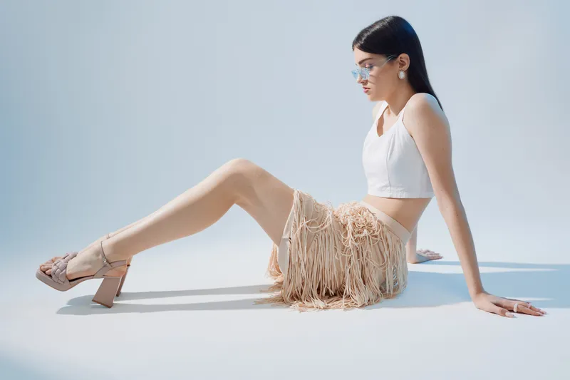 Young woman in a white crop top and beige fringe skirt poses seated on a white floor. Soft natural light highlights her fashion style and beauty concept.