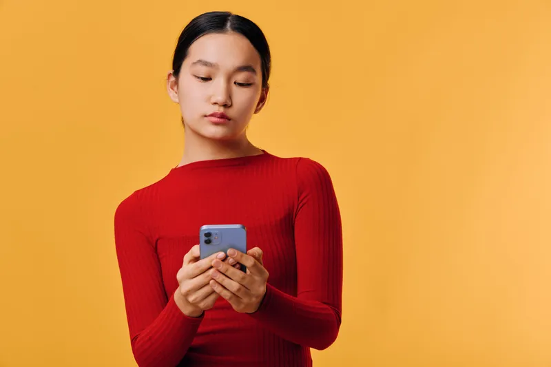 Woman in red sweater using smartphone with focused expression on yellow background. Closeup portrait of young female holding mobile device for texting and browsing content.