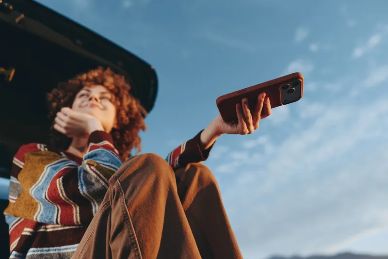 Woman lifestyle outdoors with curly hair smiling while holding smartphone recording, wearing casual striped jacket, standing near car roof, bright sky, sunny backlight, relaxed carefree moment