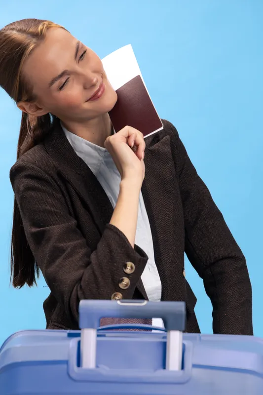 Young woman in brown jacket holding passport near blue suitcase smiles with closed eyes on blue background. Concept of travel, vacation, and tourism preparation.
