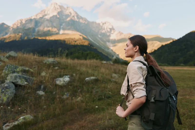 A woman with a backpack hikes through a meadow toward distant mountains, enjoying fresh air and solitude on a clear day.