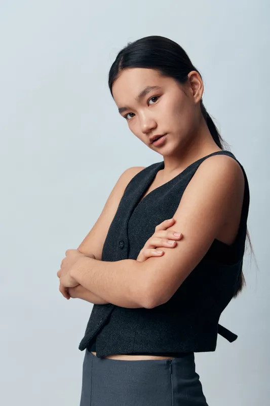 Young woman with long dark hair poses with crossed arms in black sleeveless vest and gray pants against plain light background. Confident expression and casual modern fashion style portrait.