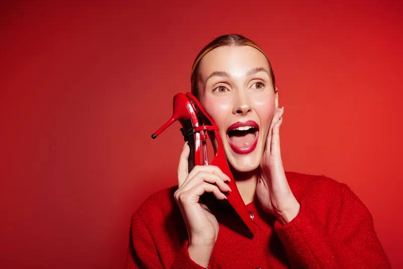 Excited woman in red sweater holds red high heel shoe near face, smiling with open mouth on red background. Concept of fashion, style, and joyful emotions.