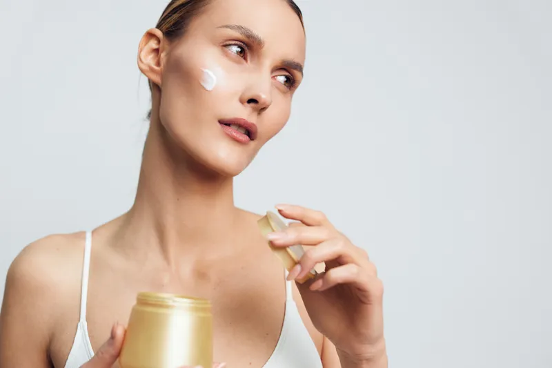 Young woman applying moisturizing cream on her face with a focused expression, isolated on a white background. Concept of skincare, beauty routine, and self-care at home.
