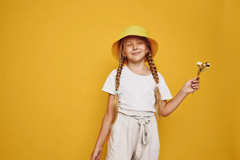 Smiling girl with braided hair wears yellow hat and white shirt holding small flowers on yellow background. Concept for childhood and happiness.