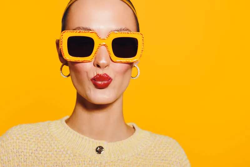 Stylish woman with bright red lipstick and large orange sunglasses poses against vibrant yellow background. Fashion concept with bold colors and confident facial expression in studio lighting.
