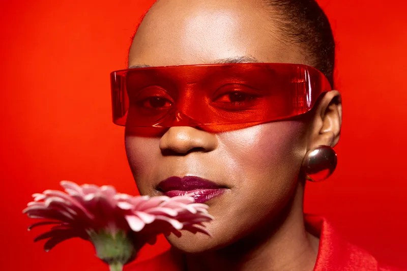 Closeup of confident black woman with dark skin wearing red sunglasses and earrings holding pink flower on red background. Concept of fashion, beauty, and elegance with vibrant colors.