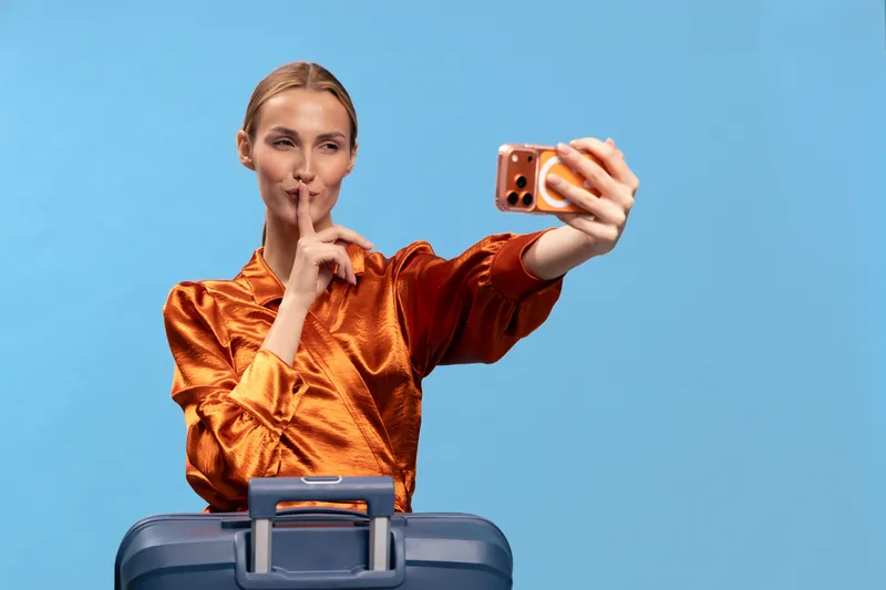 Young woman in orange satin blouse takes a selfie while holding finger to lips. Blue suitcase in front, studio background. Concept for travel secrets and quiet moments.