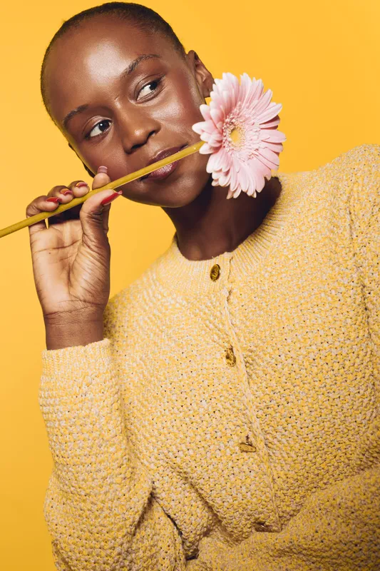 Portrait of a dark skinned African woman holding pink flower on pencil against yellow background wearing knitted sweater. Concept of beauty, nature, and positive mood.