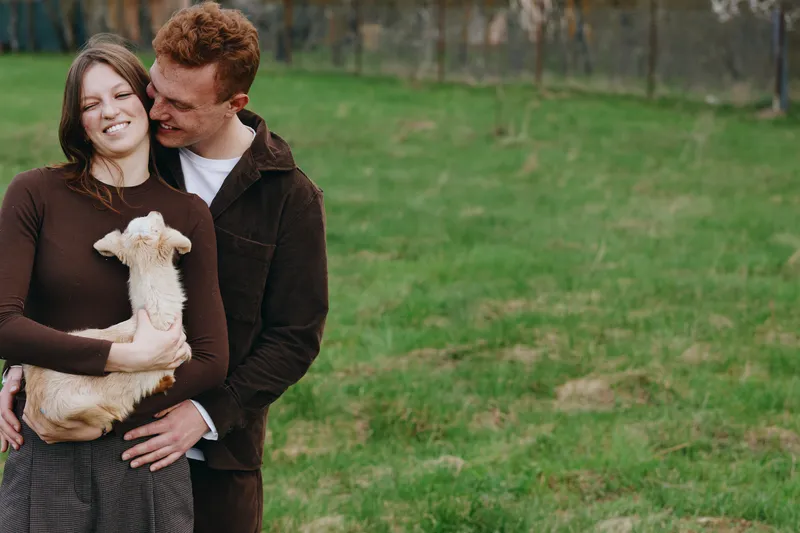 Young couple embraces outdoors while woman holds a baby goat in her arms, both smiling and enjoying a relaxed lifestyle moment in a green field.