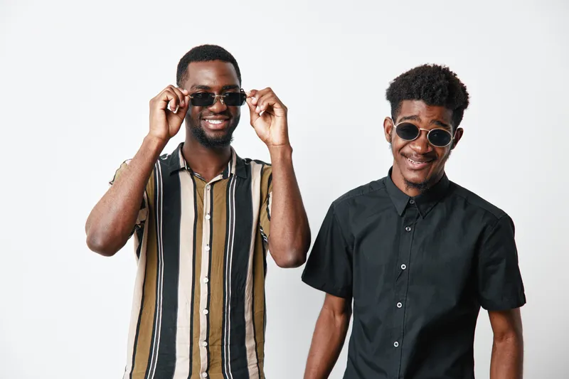 Two young black men with dark skin wearing sunglasses smiling and posing together on white background. Casual style, friendship and happiness with afro hair and modern clothes.