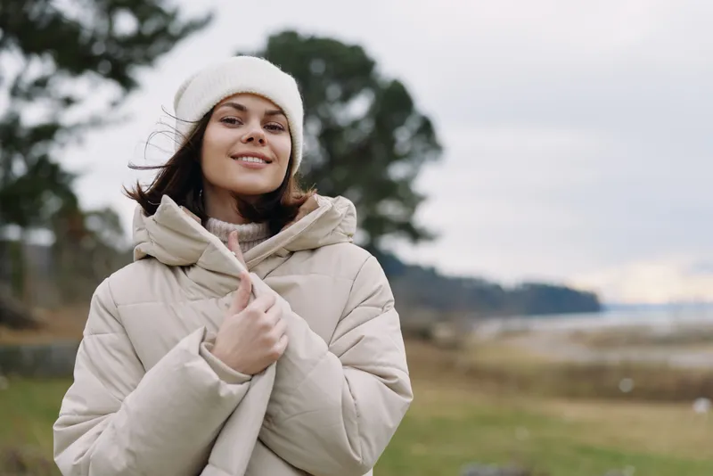 Young woman in a warm puffer coat and knit beanie enjoying a chilly day outdoors by a windswept field, smiling and relaxed.