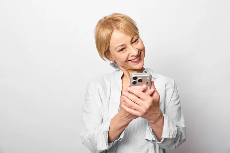 Elderly woman smiling while holding a smartphone near her face, home portrait on a bright white background, concept of technology, communication and joy today