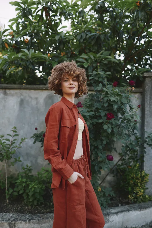Young woman with curly hair stands in a garden wearing a rust-colored suit, posing with hands in pockets and a calm confident smile, while soft natural light highlights fabric texture and greenery