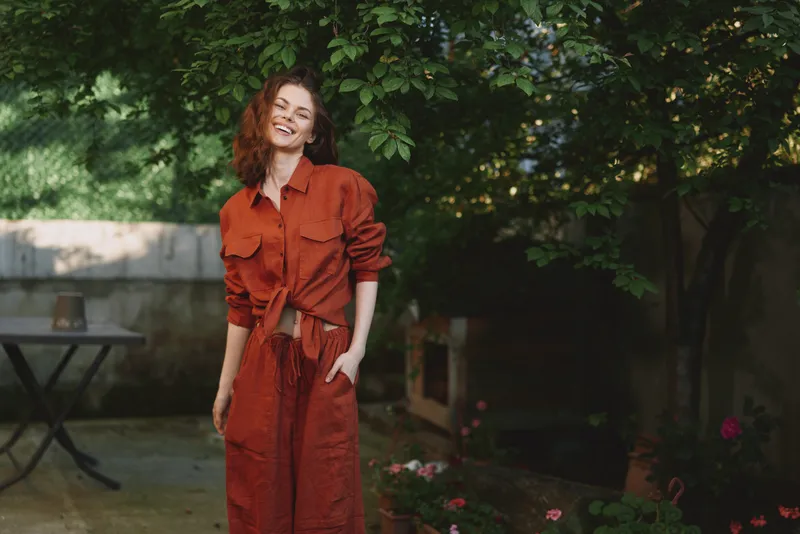 Vibrant woman in a burnt orange dress stands in a lush garden, smiling softly, capturing a casual lifestyle moment outdoors with natural light and relaxed pose