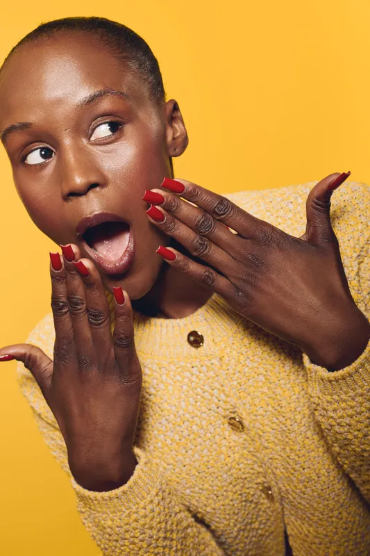 Surprised African woman with dark skin and red nails wearing yellow knitted sweater looks away with open mouth on yellow background. Concept of emotions, beauty, fashion, and expression.