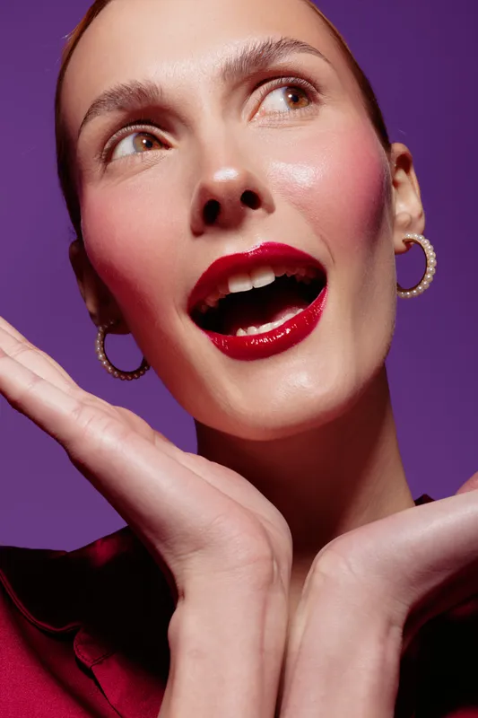 Close-up of a smiling woman with glossy red lips and hoop earrings posing with hands near face on purple background. Concept of beauty, makeup, fashion, and joyful expression.