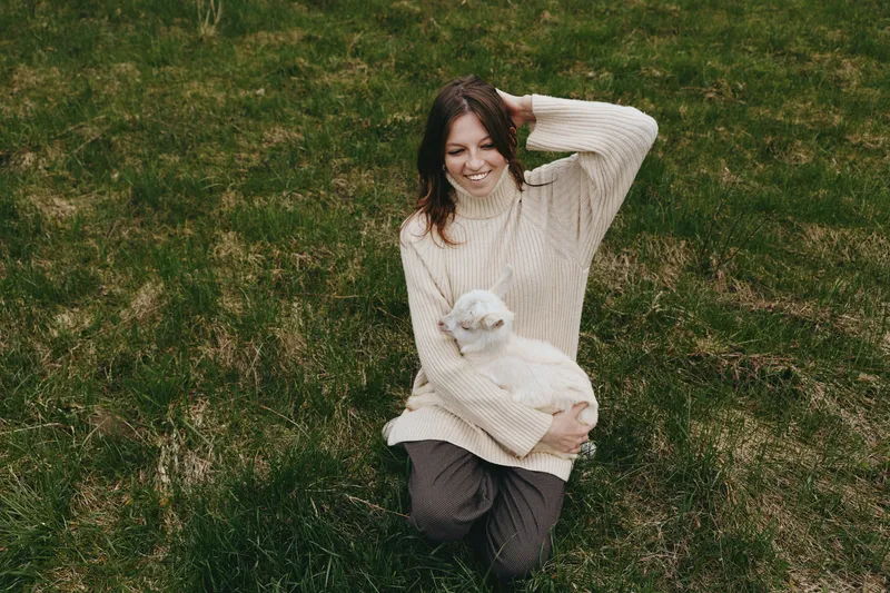Young woman with long brown hair smiles while sitting on grass holding a small white baby goat in her arms during a relaxed outdoor lifestyle moment.