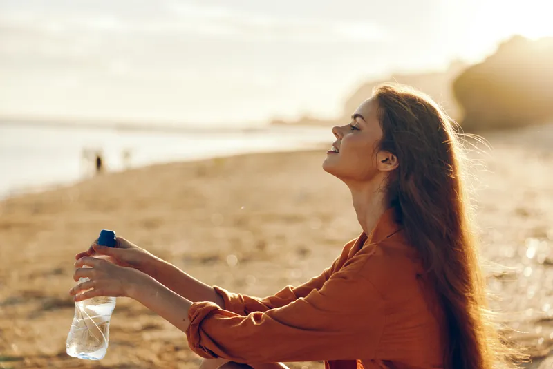 Woman on beach at sunset drinks water from a bottle, staying calm for hydration and recovery from dehydration, easing thirst in sunlight during summer outdoors wellness