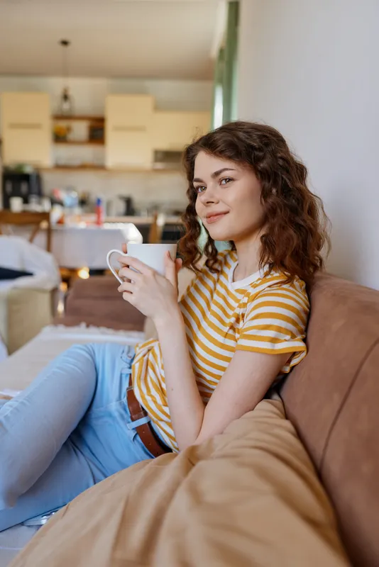 Casual woman relaxes on a sofa at home, holding a mug, wearing a striped yellow shirt and jeans, with soft natural light in a cozy living room and open kitchen background.