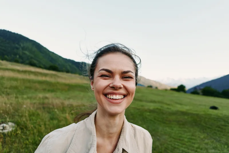 Joyful woman outdoors, smiling in a sunlit field, relaxed pose, natural colors, lush grass, distant mountains, fresh air, candid moment, warm breeze